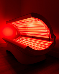 A red light therapy device or tanning bed glowing brightly with intense red light, set against a dark red and black background.